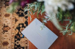 A quiet scene of a light grey gest book on a wooden table, a patterned carpet in the background, and foliage in the foreground