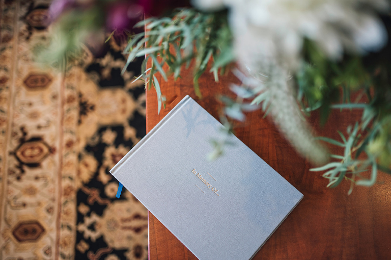 A quiet scene of a light grey gest book on a wooden table, a patterned carpet in the background, and foliage in the foreground